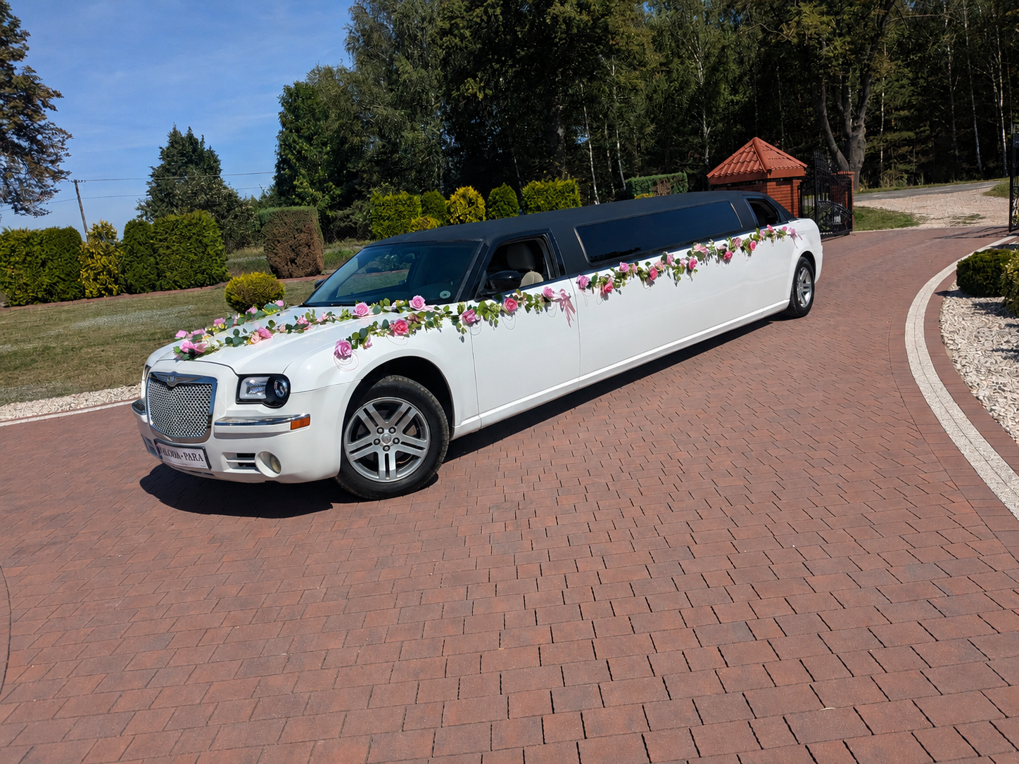 Biała limuzyna Chrysler z girlandą róż i tablicą MŁODA-PARA na brukowanym podjeździe