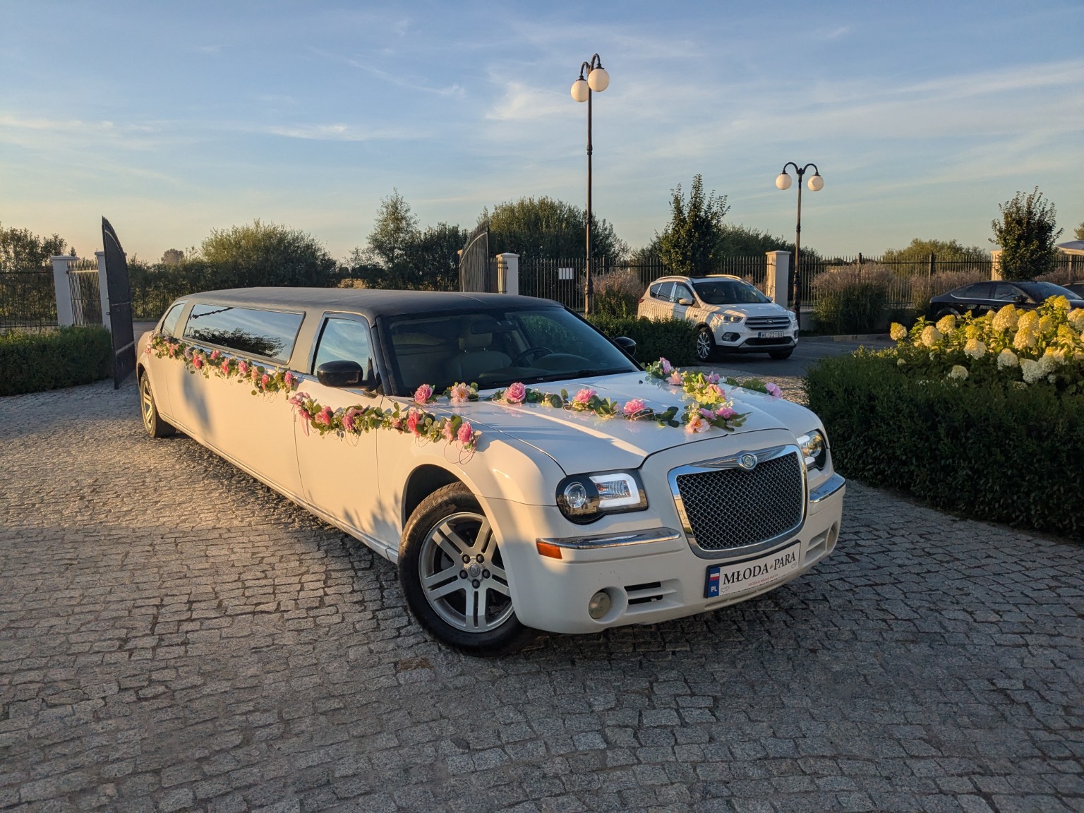 Biała limuzyna Chrysler z różową girlandą kwiatów i tablicą MŁODA-PARA na brukowanym podjeździe